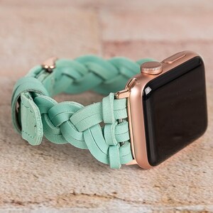May include: A rose gold Apple Watch with a mint green braided leather band. The watch face is black. The band is made of interwoven leather strips and has a buckle closure. The watch is displayed on a light brown surface.