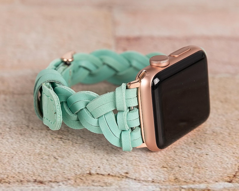 May include: A rose gold Apple Watch with a mint green braided leather band. The watch face is black. The band is made of interwoven leather strips and has a buckle closure. The watch is displayed on a light brown surface.