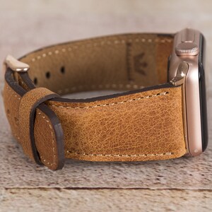 May include: A brown leather watch strap with a rose gold Apple Watch attached. The strap has a textured finish and is stitched with white thread.