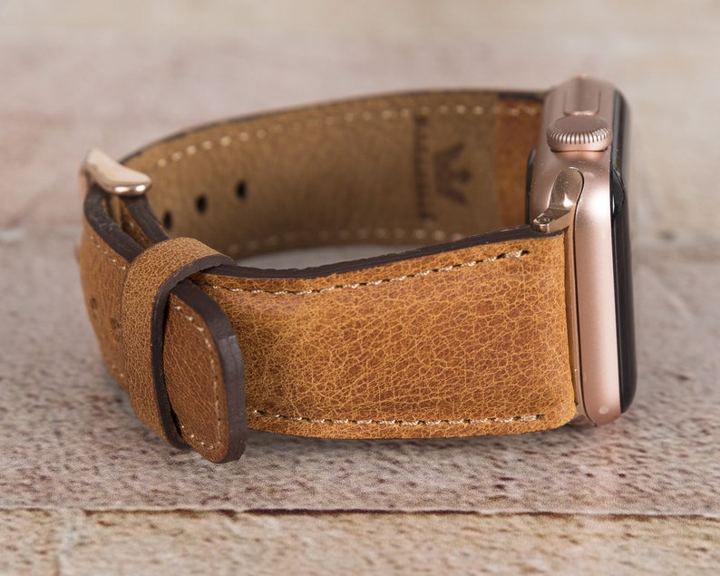 May include: A brown leather watch strap with a rose gold Apple Watch attached. The strap has a textured finish and is stitched with white thread.