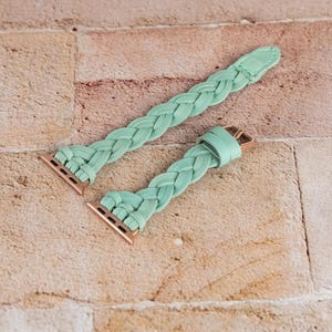 May include: Two mint green braided watch bands with rose gold connectors and clasps. The bands are made of a soft, woven material and are displayed on a light brown brick surface. The bands are designed to fit a smartwatch.
