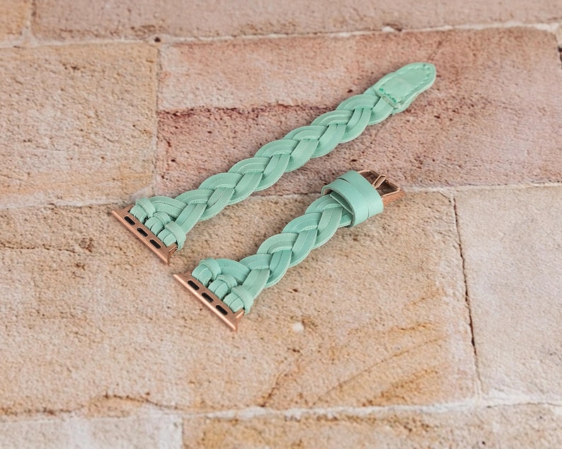 May include: Two mint green braided watch bands with rose gold connectors and clasps. The bands are made of a soft, woven material and are displayed on a light brown brick surface. The bands are designed to fit a smartwatch.