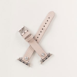 Pode incluir: Uma pulseira de rel&oacute;gio de couro bege claro com detalhes prateados e strass transparentes. A pulseira &eacute; projetada para um Apple Watch.