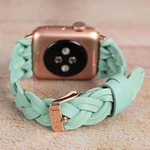 May include: Rose gold Apple Watch with a braided mint green leather band. The band features a gold buckle. The watch is displayed on a light brown surface.