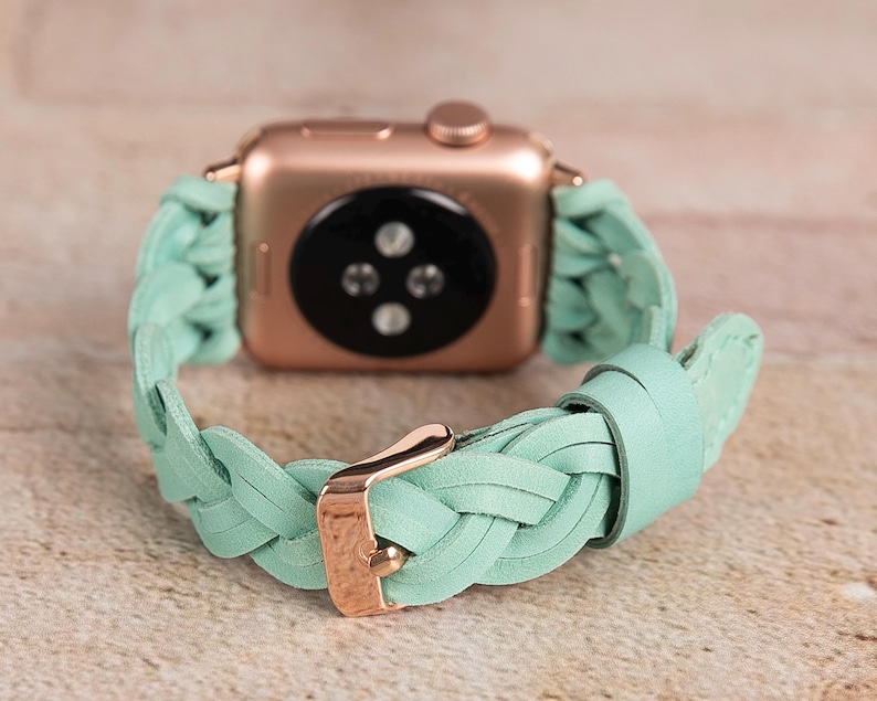 May include: Rose gold Apple Watch with a braided mint green leather band. The band features a gold buckle. The watch is displayed on a light brown surface.