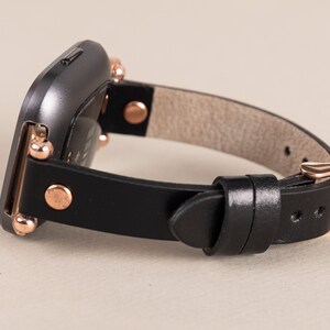May include: A black leather watch band with rose gold accents and a buckle closure. The band is designed for a square-shaped smartwatch.