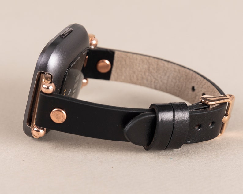 May include: A black leather watch band with rose gold accents and a buckle closure. The band is designed for a square-shaped smartwatch.