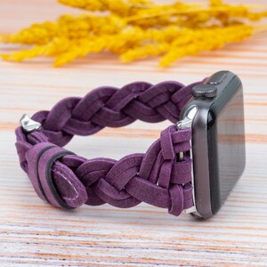 Pu&ograve; includere: Un cinturino in pelle intrecciata viola per Apple Watch. Il cinturino &egrave; realizzato in pelle morbida e flessibile e presenta un design intrecciato. Il cinturino &egrave; mostrato su una superficie in legno chiaro.