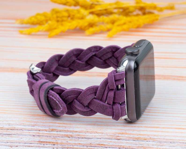 Pu&ograve; includere: Un cinturino in pelle intrecciata viola per Apple Watch. Il cinturino &egrave; realizzato in pelle morbida e flessibile e presenta un design intrecciato. Il cinturino &egrave; mostrato su una superficie in legno chiaro.