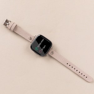 Puede incluir: Una pulsera de reloj inteligente de cuero rosa claro con una esfera cuadrada negra que muestra 12.000 pasos y 67 ppm.