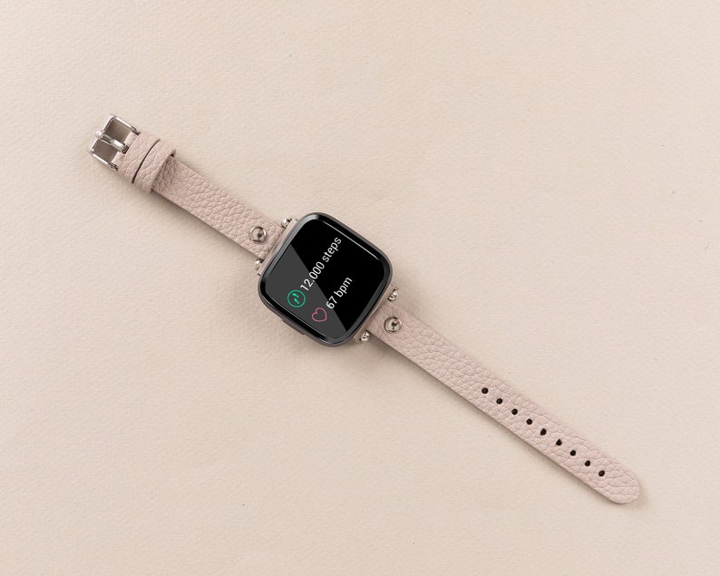 Puede incluir: Una pulsera de reloj inteligente de cuero rosa claro con una esfera cuadrada negra que muestra 12.000 pasos y 67 ppm.