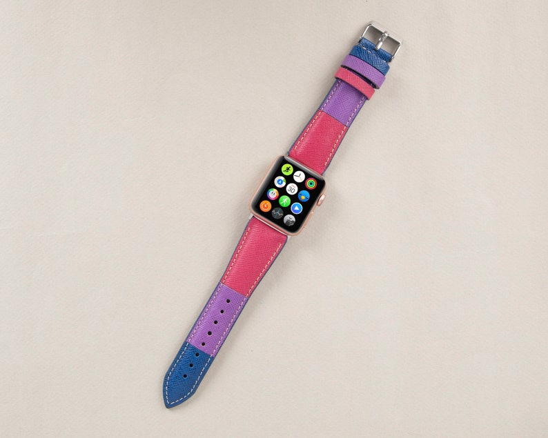 Puede incluir: Una banda de reloj Apple de cuero rosa, morado y azul con una esfera de reloj Apple dorada.