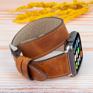 K&ouml;nnte beinhalten: Ein braunes Lederarmband mit einer silbernen Schlie&szlig;e. Das Armband ist um eine Smartwatch mit schwarzem Zifferblatt und bunten Symbolen gewickelt.