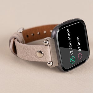 Puede incluir: Un rastreador de fitness negro con una banda de cuero gris. La pantalla muestra "12.000 pasos" y "67 ppm".