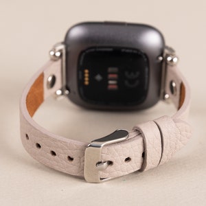 Puede incluir: Una correa de reloj de cuero gris claro con herrajes plateados y una esfera de reloj inteligente negra. La esfera del reloj tiene un peque&ntilde;o punto blanco y tres l&iacute;neas blancas.