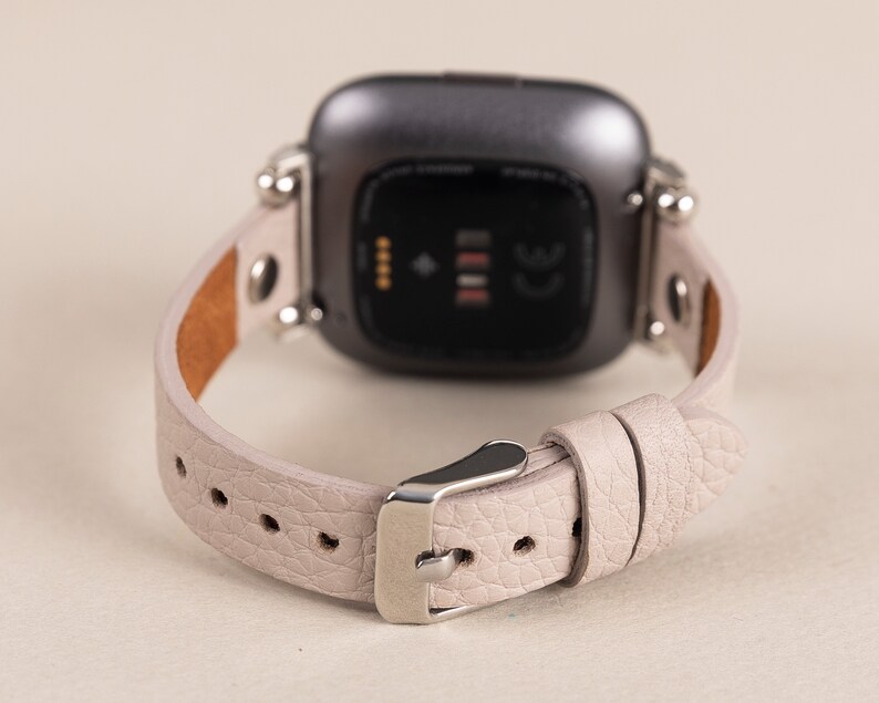 Puede incluir: Una correa de reloj de cuero gris claro con herrajes plateados y una esfera de reloj inteligente negra. La esfera del reloj tiene un peque&ntilde;o punto blanco y tres l&iacute;neas blancas.