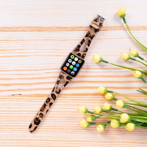 May include: A leopard print Apple Watch band with a gold buckle. The watch face is visible and shows a variety of apps. The band is lying on a light wood surface with yellow flowers.