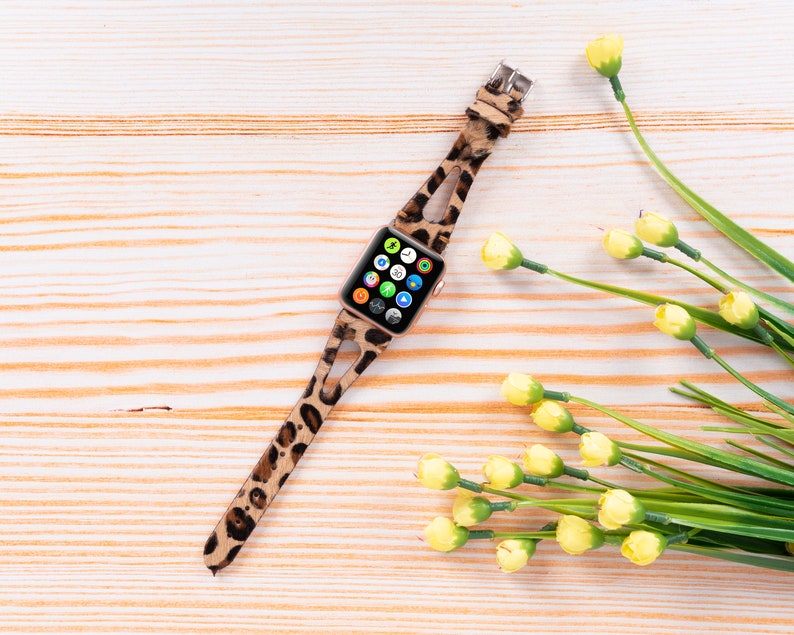 May include: A leopard print Apple Watch band with a gold buckle. The watch face is visible and shows a variety of apps. The band is lying on a light wood surface with yellow flowers.