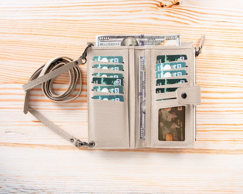 Pu&ograve; includere: Un portafoglio in pelle beige testurizzata con tracolla staccabile, dotato di molteplici scomparti per carte, una finestra trasparente per documenti e uno scomparto con cerniera. Il portafoglio &egrave; aperto e contiene diverse carte di credito verdi.