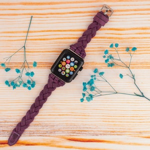 Pu&ograve; includere: Un cinturino in pelle intrecciata viola per un Apple Watch. Il quadrante dell'orologio &egrave; visibile e mostra l'ora e diverse icone di app.