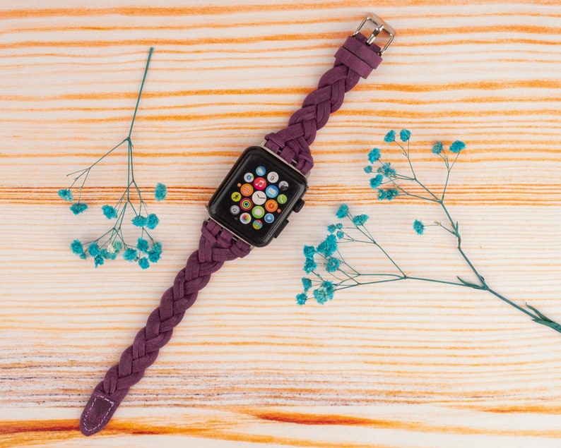 Pu&ograve; includere: Un cinturino in pelle intrecciata viola per un Apple Watch. Il quadrante dell'orologio &egrave; visibile e mostra l'ora e diverse icone di app.