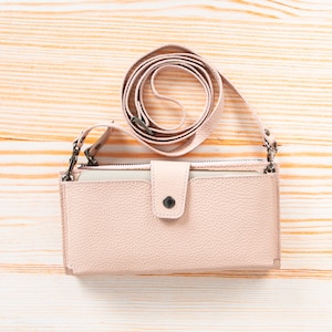 Pu&ograve; includere: Un portafoglio a tracolla color rosa pallido con texture granulata di forma rettangolare, dotato di chiusura centrale con bottone a pressione, scomparto superiore con cerniera e tracolla regolabile abbinata con attacchi in metallo argentato.