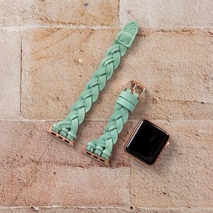 May include: A mint green braided leather watch band with a gold-colored buckle and connectors. A rose gold smartwatch with a black screen is next to the band. The watch band is shown on a brick background.