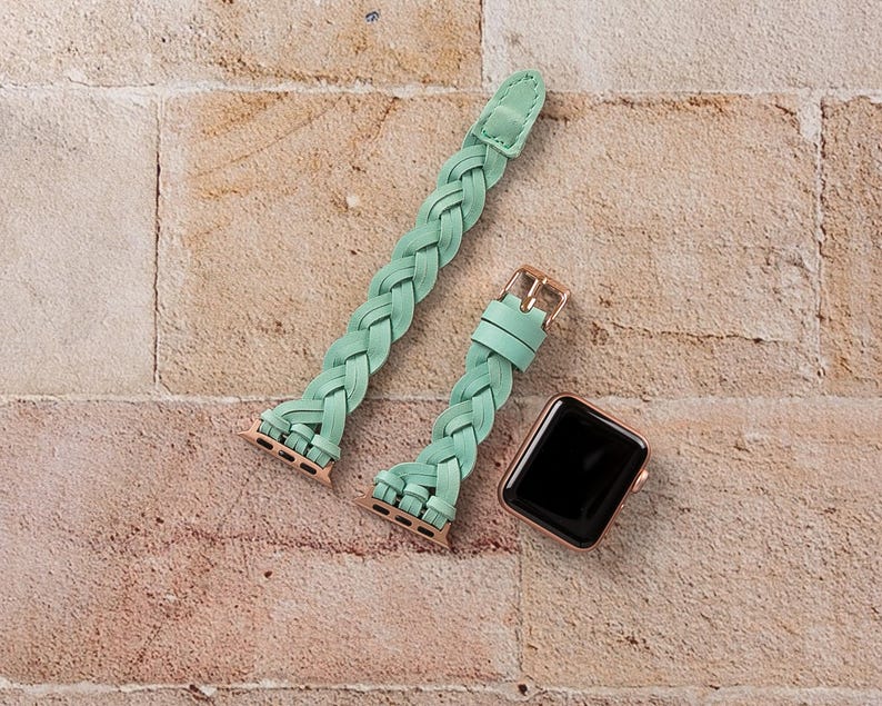 May include: A mint green braided leather watch band with a gold-colored buckle and connectors. A rose gold smartwatch with a black screen is next to the band. The watch band is shown on a brick background.