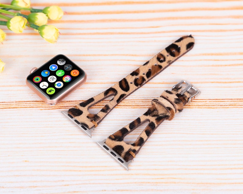 May include: Two leopard print Apple Watch bands with a cutout design. The bands are brown and tan with black spots. The bands are shown on a light wood surface.