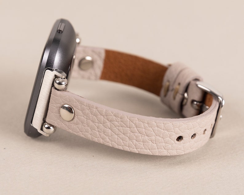 Puede incluir: Una correa de reloj de cuero gris claro con detalles plateados y cierre de hebilla.
