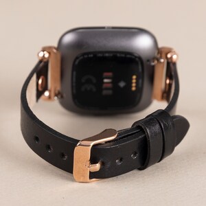 May include: A black leather watch band with a rose gold buckle and a black and silver smartwatch face. The watch face has a CE mark and a small diamond symbol.