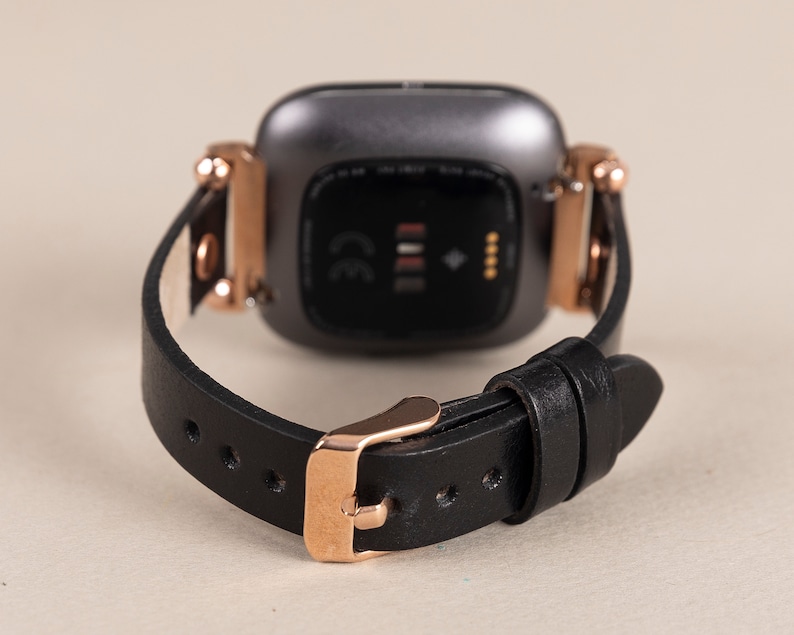 May include: A black leather watch band with a rose gold buckle and a black and silver smartwatch face. The watch face has a CE mark and a small diamond symbol.