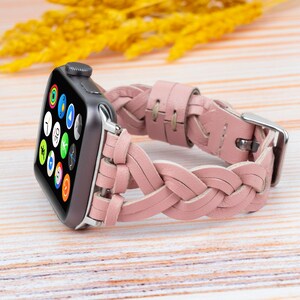 K&ouml;nnte beinhalten: Ein rosafarbenes geflochtenes Lederarmband f&uuml;r eine Apple Watch. Das Armband ist aus weichem, geschmeidigem Leder gefertigt und hat ein geflochtenes Design. Das Armband wird auf einer hellen Holzoberfl&auml;che gezeigt.