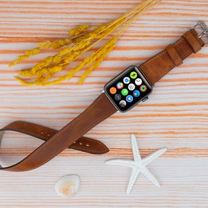 K&ouml;nnte beinhalten: Ein braunes Lederarmband f&uuml;r eine Apple Watch. Das Armband ist auf einem hellen Holzhintergrund mit einem Seestern und Muscheln abgebildet.