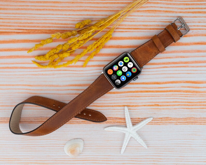 K&ouml;nnte beinhalten: Ein braunes Lederarmband f&uuml;r eine Apple Watch. Das Armband ist auf einem hellen Holzhintergrund mit einem Seestern und Muscheln abgebildet.