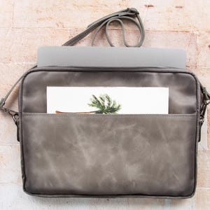 May include: Gray leather messenger bag with a shoulder strap. The bag has a front pocket and a top zipper closure. A laptop is inside the bag, and a watercolor painting of a palm tree is in the front pocket.