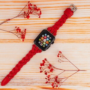 Puede incluir: Una banda de reloj Apple trenzada roja con una esfera de reloj Apple negra. El reloj est&aacute; sobre una superficie de madera con flores rojas.