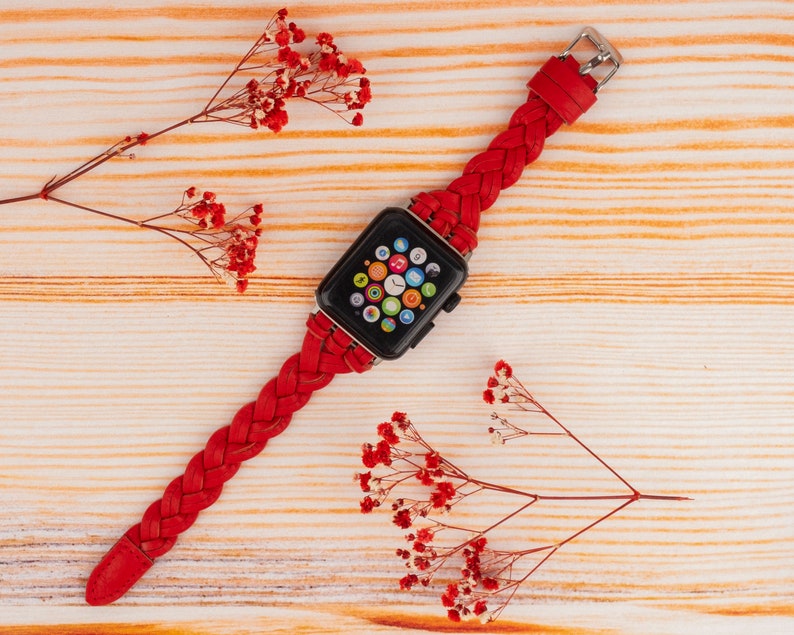 Puede incluir: Una banda de reloj Apple trenzada roja con una esfera de reloj Apple negra. El reloj est&aacute; sobre una superficie de madera con flores rojas.