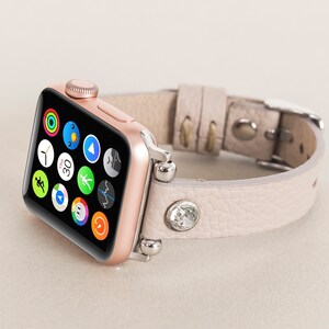 Pode incluir: Um Apple Watch dourado rosa com uma pulseira de couro bege claro adornada com um cristal. O mostrador do rel&oacute;gio exibe uma variedade de &iacute;cones de aplicativos.