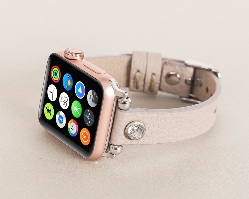Pode incluir: Um Apple Watch dourado rosa com uma pulseira de couro bege claro adornada com um cristal. O mostrador do rel&oacute;gio exibe uma variedade de &iacute;cones de aplicativos.