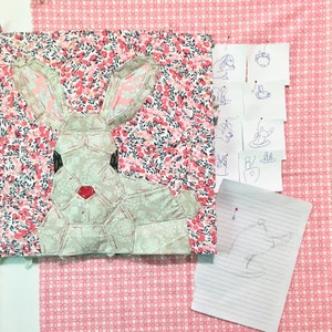 May include: A patchwork bunny design with a red gem nose, set on a floral fabric. The bunny is made from geometric shapes in neutral colours. Sketches of bunnies are pinned nearby, indicating a craft project.
