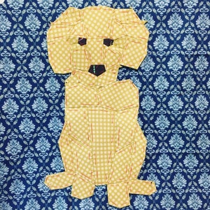May include: A yellow fabric dog with white polka dots, sewn together in a patchwork style. The dog is standing on a blue and white patterned fabric background.