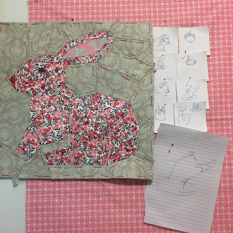 May include: A patchwork quilt block featuring a pink and white floral bunny rabbit design. The block is surrounded by a green and white patterned fabric. There are several small pieces of paper with hand-drawn bunny sketches pinned to the fabric.