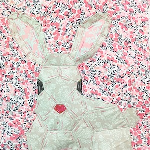 May include: A patchwork quilt block featuring a white bunny with pink ears and a red nose. The bunny is surrounded by a pink and white floral pattern.