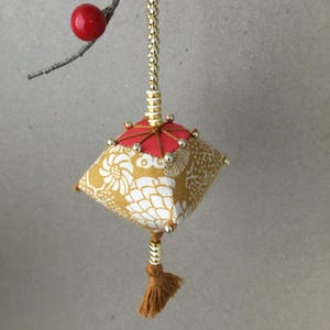 May include: A handmade, decorative ornament with a square, patterned body in gold and white floral fabric. It features a red top, gold bead accents, and a brown tassel. The ornament hangs from a gold beaded chain and a small branch with a red berry.