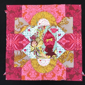 May include: A colorful patchwork quilt block with a central image of a stylized figure with pink hair and a bow. The block is made up of various fabrics with floral and geometric patterns in shades of pink, red, yellow, and green.