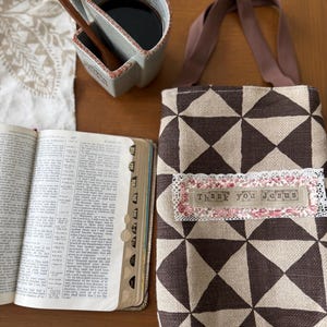 May include: A brown and cream geometric patterned tote bag with brown handles. The bag has a decorative label that reads "Thank you Jesus". A ceramic mug with coffee and a wooden spoon is next to the bag. An open book is also in the image.