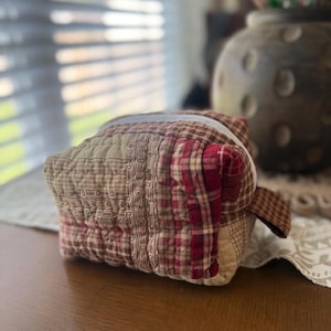 May include: A rectangular quilted pouch featuring a patchwork design of red, beige, and brown fabrics. The exterior displays a mix of plaid, striped, and solid patterns with visible stitching throughout. A small fabric tab extends from the side.