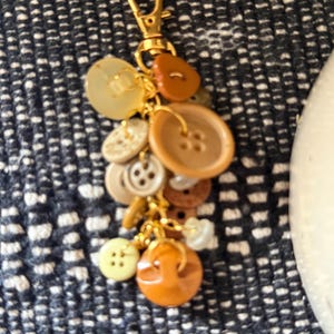 Peut inclure: Un porte-clés composé d'une collection de boutons assortis dans diverses nuances de marron, beige, crème et blanc cassé. Les boutons sont fixés à un fermoir mousqueton doré par de petits anneaux dorés, créant un design de breloque en grappe.