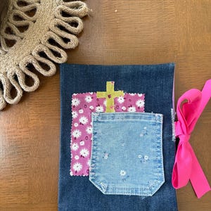 May include: A handcrafted denim book cover featuring a pink floral patch and a light blue denim pocket. A yellow cross is appliquéd on the pink patch. A bright pink ribbon is tied to the side.